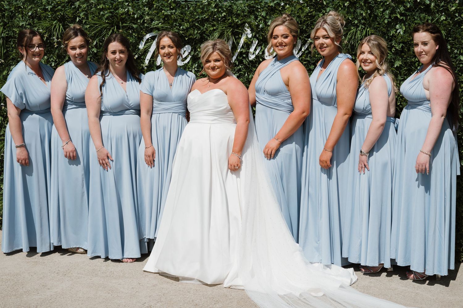 Bride in white gown poses with eight bridesmaids in matching light blue dresses outdoors.
