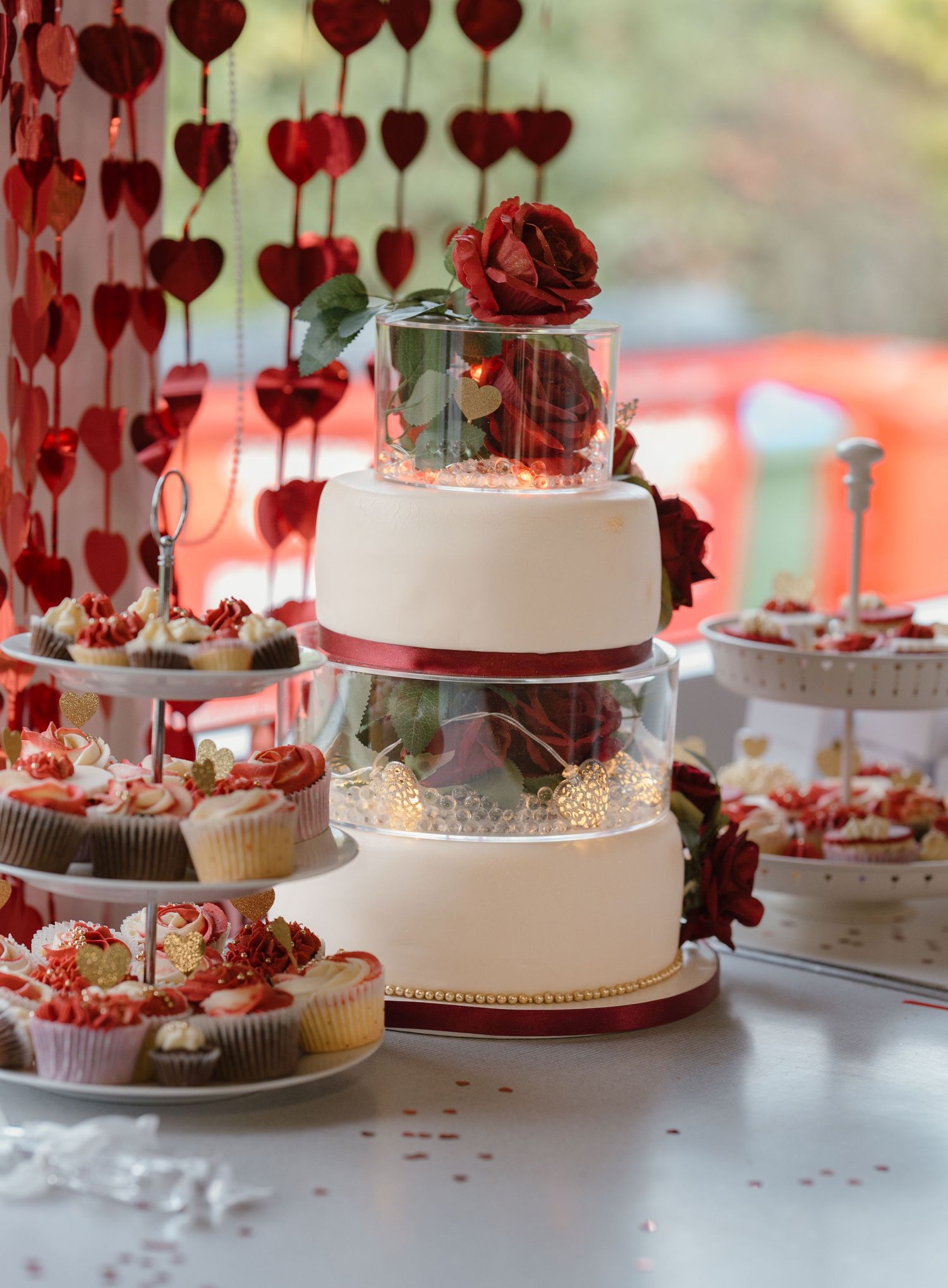 Elegant three-tier white wedding cake with red roses and ribbon, surrounded by cupcakes and heart decorations.
