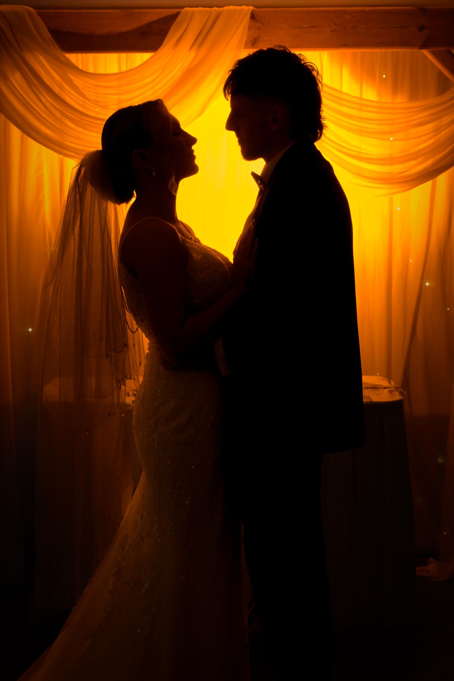 Silhouettes of a couple in formal wedding attire stand against warm orange backlit drapes in a romantic series of poses. Sitwell Arms Sheff