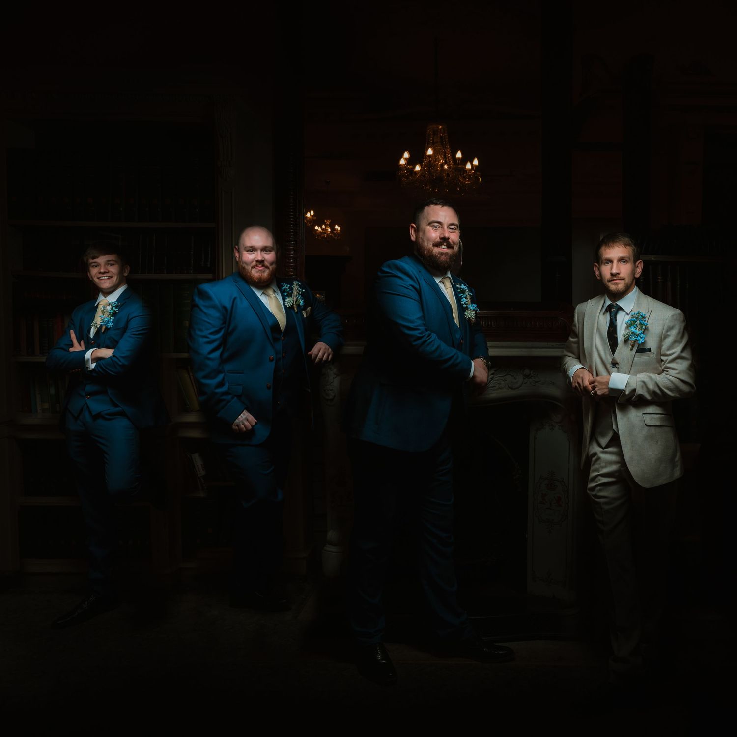 Four groomsmen in formal suits pose in a dark room with elegant chandelier lighting in the background. Venue at Wortley Hall