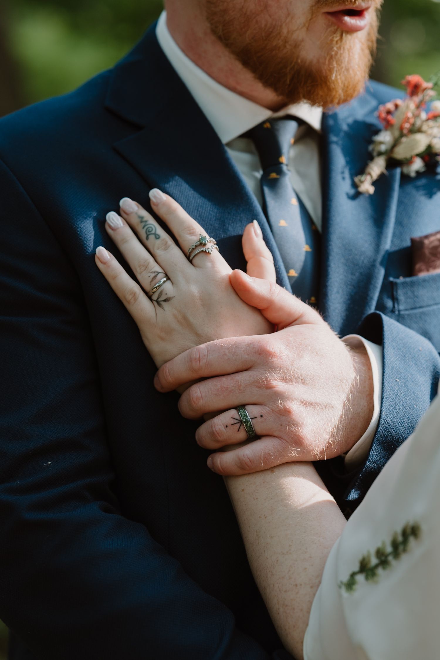 Close-up of bride and groom hands showing wedding rings and floral tattoos, groom in navy suit with boutonniere.