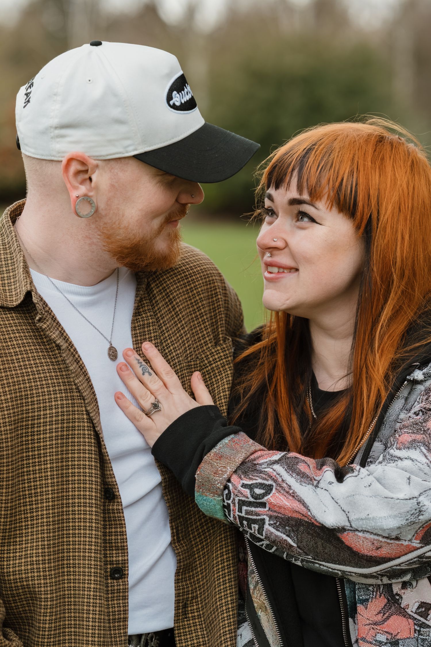 A smiling redhead woman and a man in a white cap share a tender moment outdoors in a lush green park.