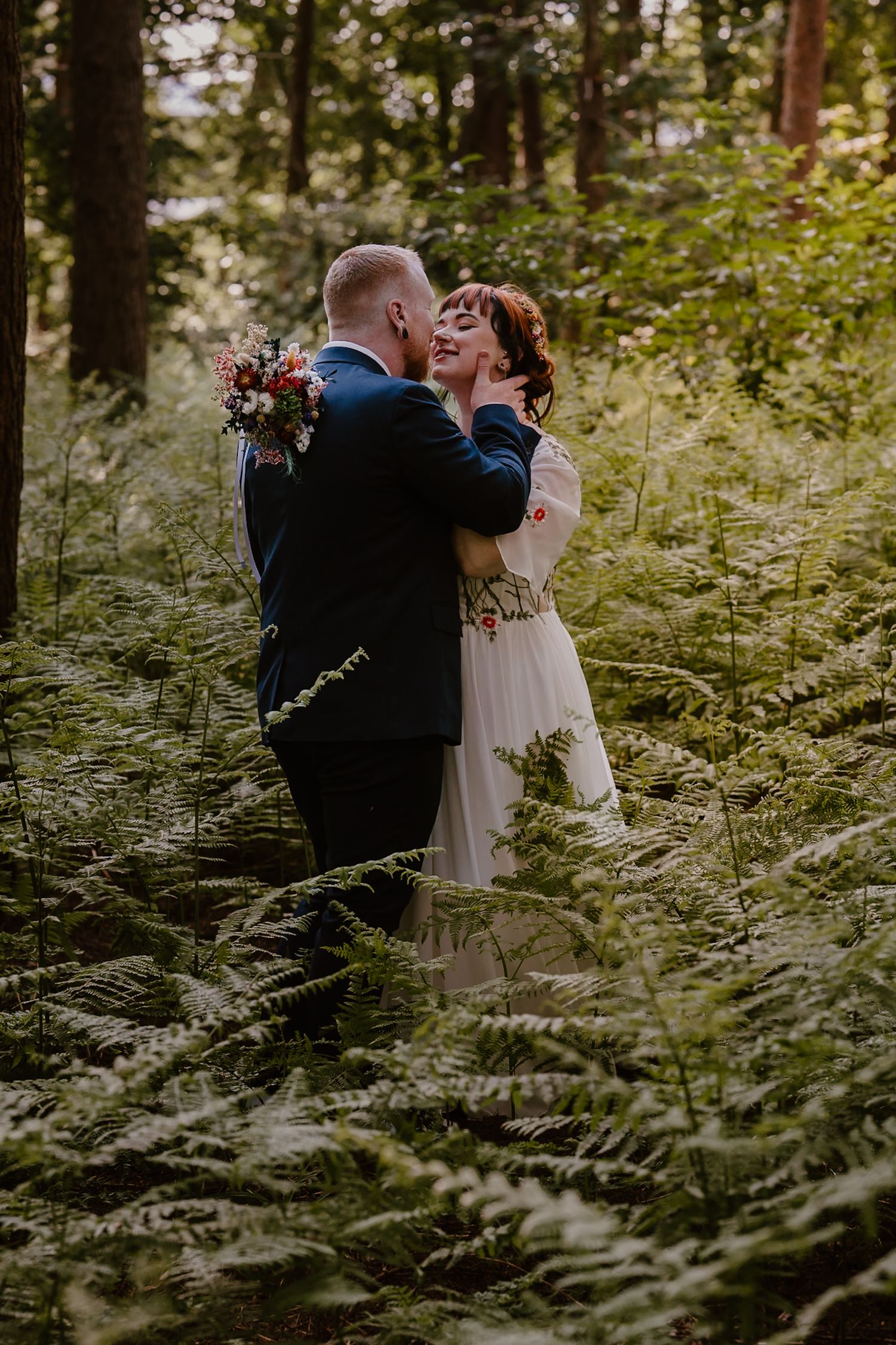Bride and groom embrace romantically among lush forest ferns, bride in white gown holding colorful bouquet.
