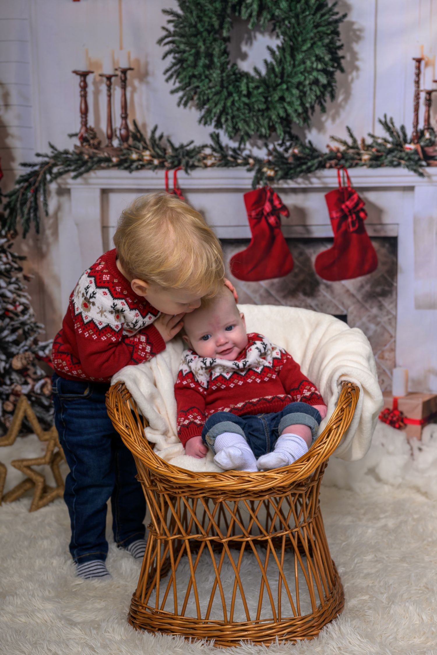 Two young brothers in matching red Christmas sweaters pose by a decorated fireplace with stockings and a wreath.