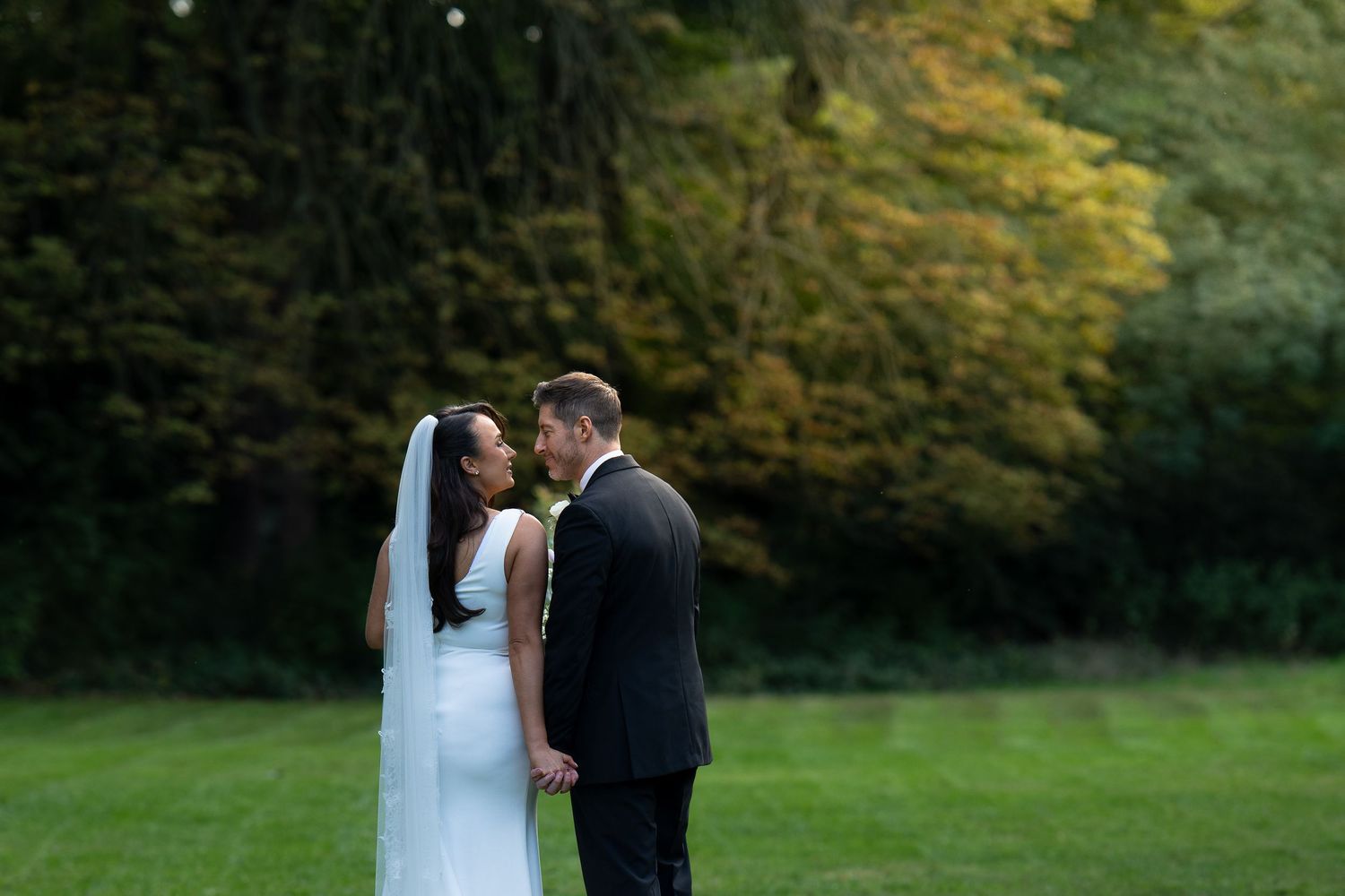 A bride and groom share a romantic moment on a lush green lawn with autumn trees in the background.