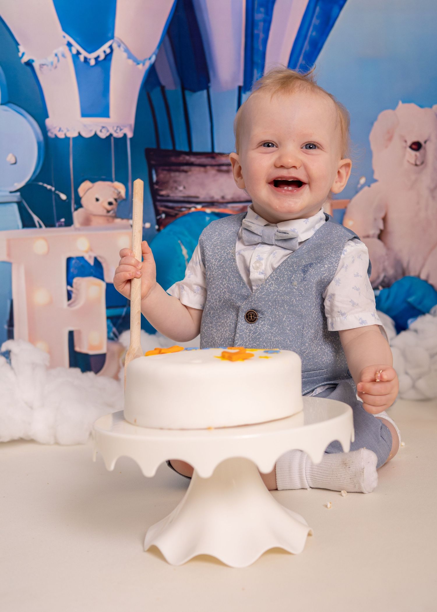Cake Smash - A baby wearing a grey outfit sits at a white table with a cake and paper snowflake decorations in a blue-themed setting.