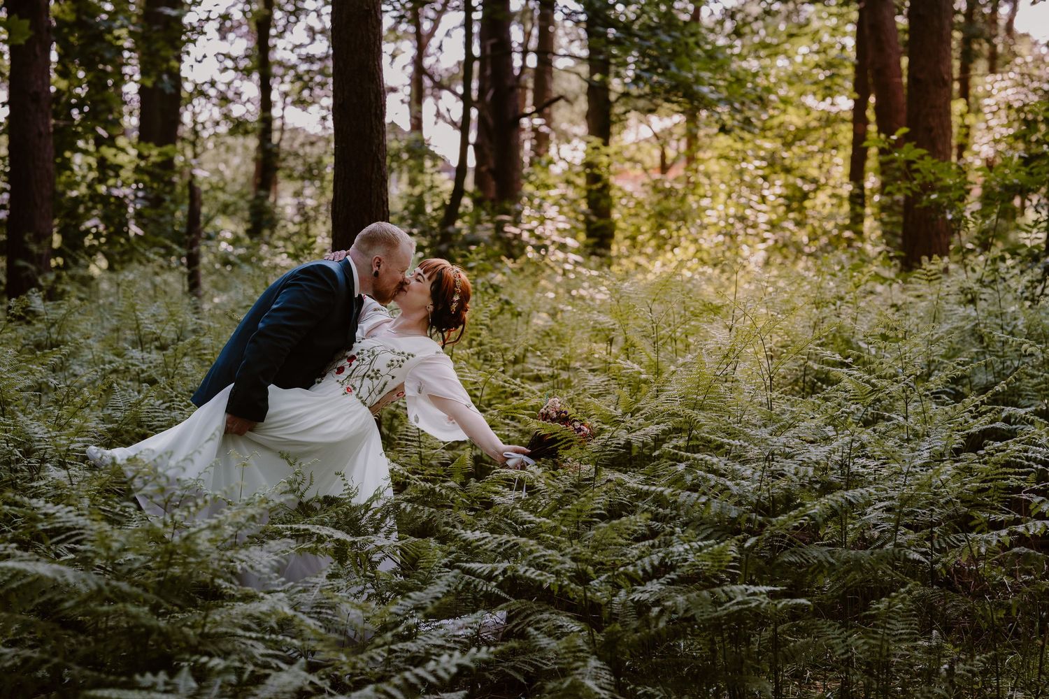 Bride and groom share a romantic dip kiss surrounded by lush green ferns in a sunlit forest wedding photo.
