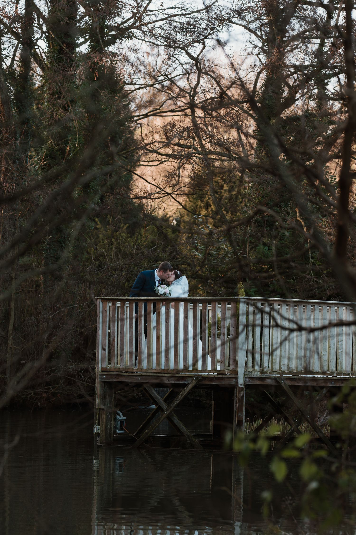 A couple sits on a wooden dock overlooking a tranquil pond surrounded by trees at sunset. Whitley Hall Sheffield