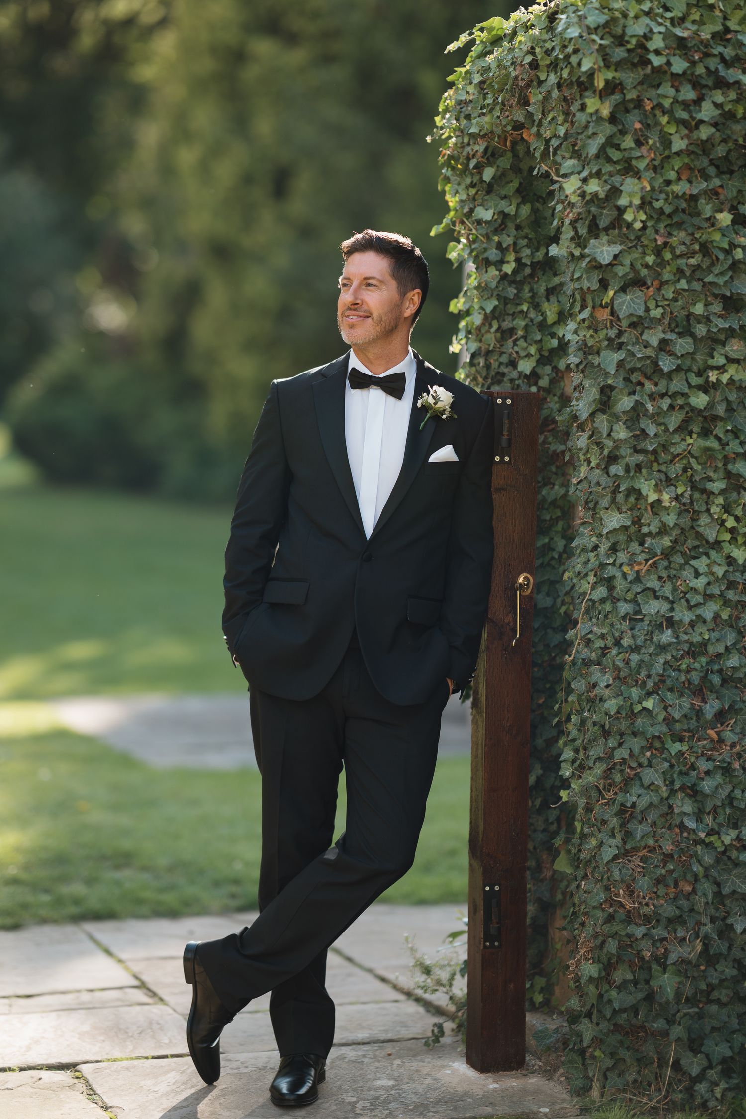Groom in black tuxedo with bow tie and boutonniere leans against ivy-covered wall in sunlit garden.