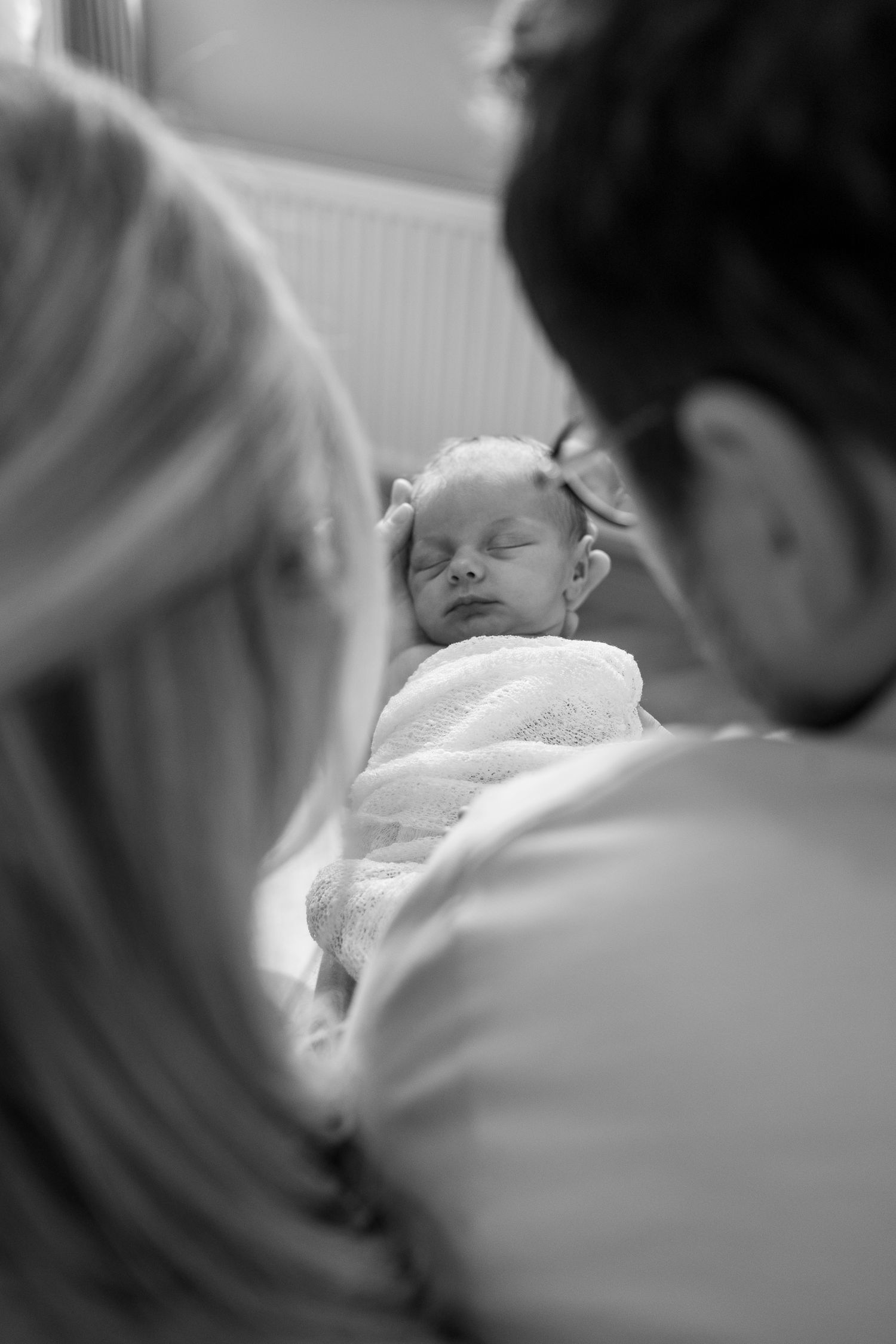 Black and white photo of parents gazing lovingly at their sleeping newborn baby wrapped in a white blanket.