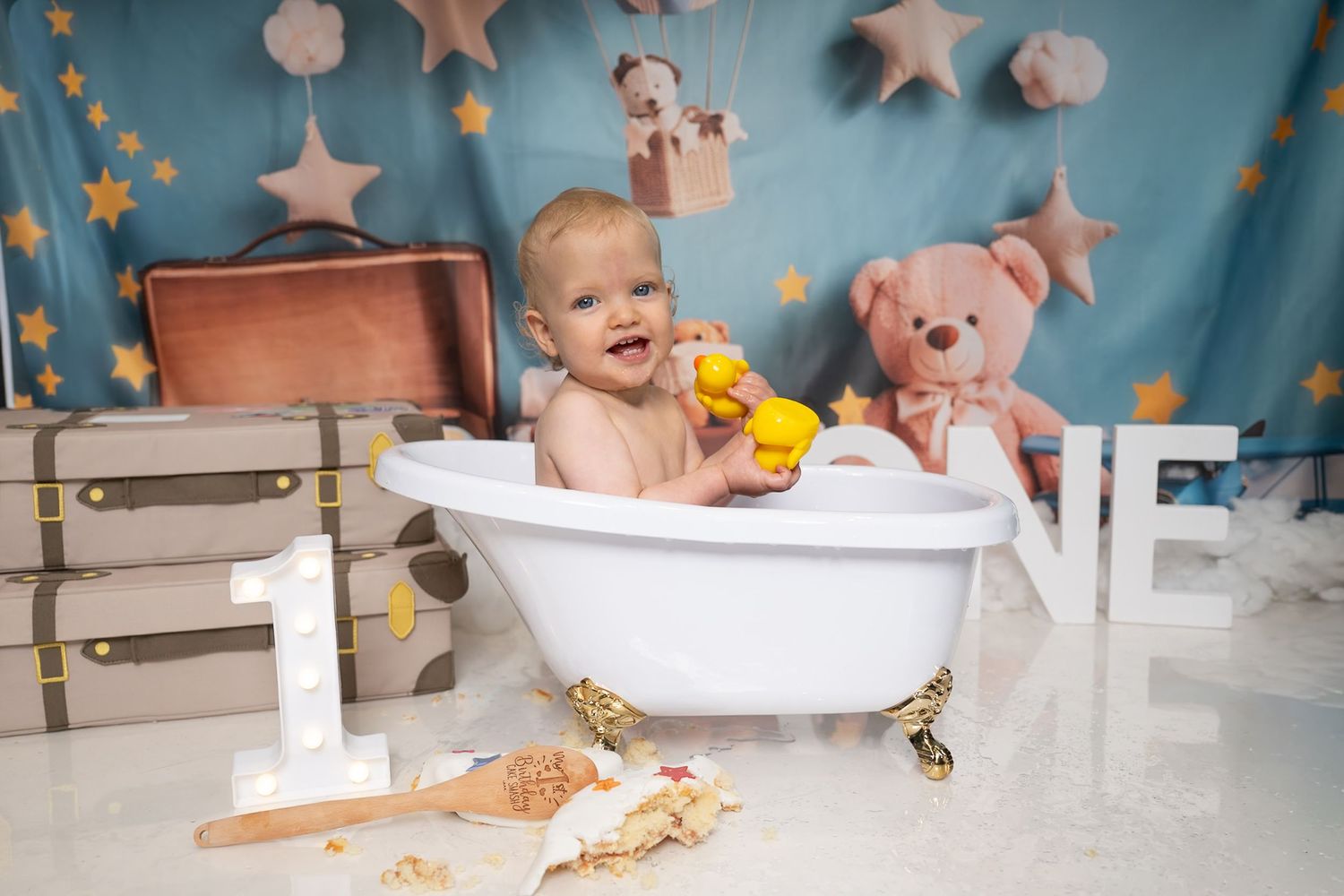 Baby smiling in a white bathtub with rubber ducks, teddy bear, and star decorations during a first birthday cake smash photo shoot.