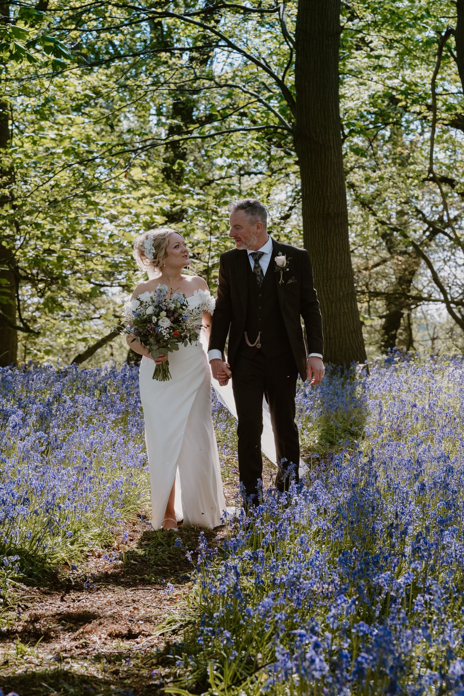 A bride and groom walk hand-in-hand through a bluebell woodland, sharing a loving glance on their wedding day.