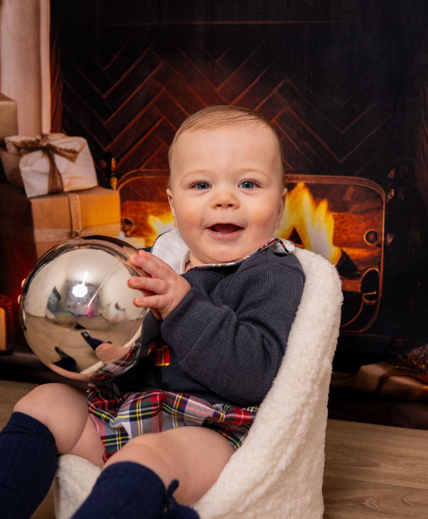 Christmas Photoshoot - Baby in plaid outfit sits in white chair holding silver ornament ball with cozy fireplace glowing in background.