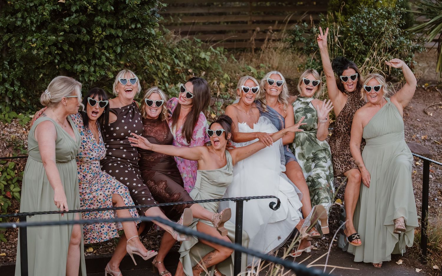 A joyful bride poses with her bridesmaids in colorful dresses and sunglasses outdoors at a wedding celebration.