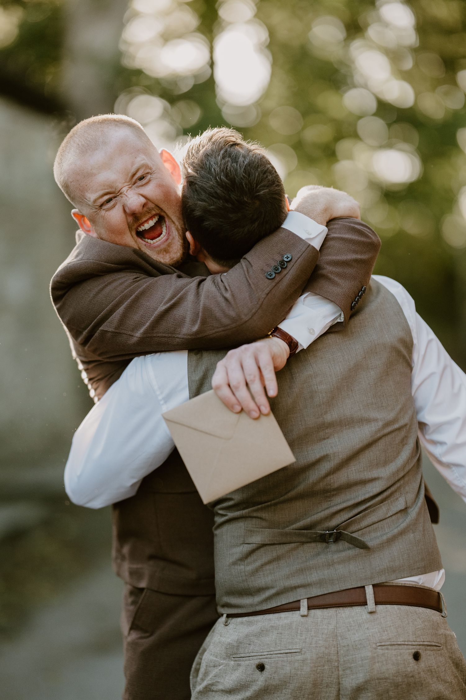 Two men in brown suits share an emotional hug at a wedding, one laughing joyfully while holding an envelope.