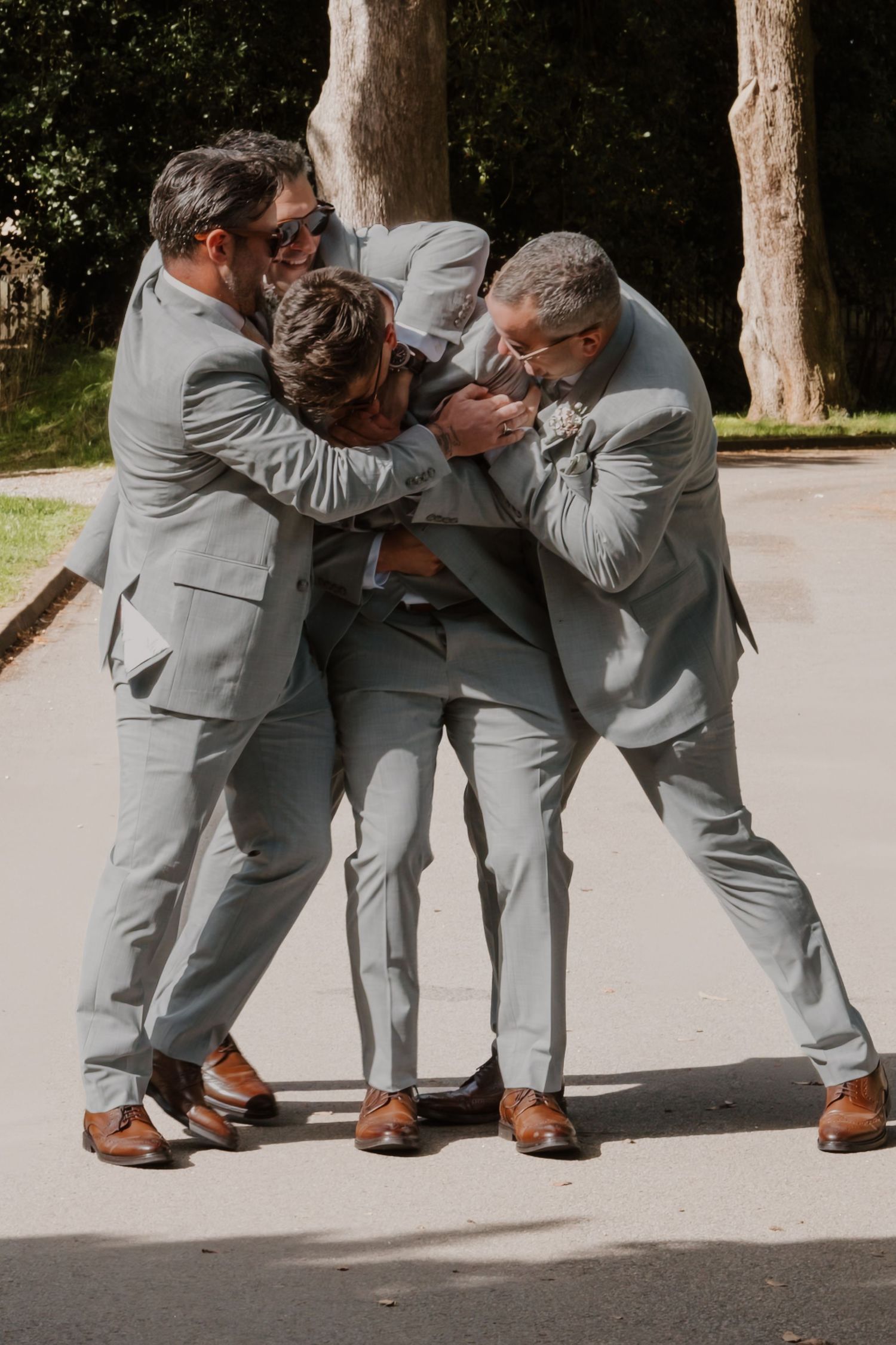 Three groomsmen in matching light gray suits and brown shoes playfully hugging outdoors on a sunny wedding day.
