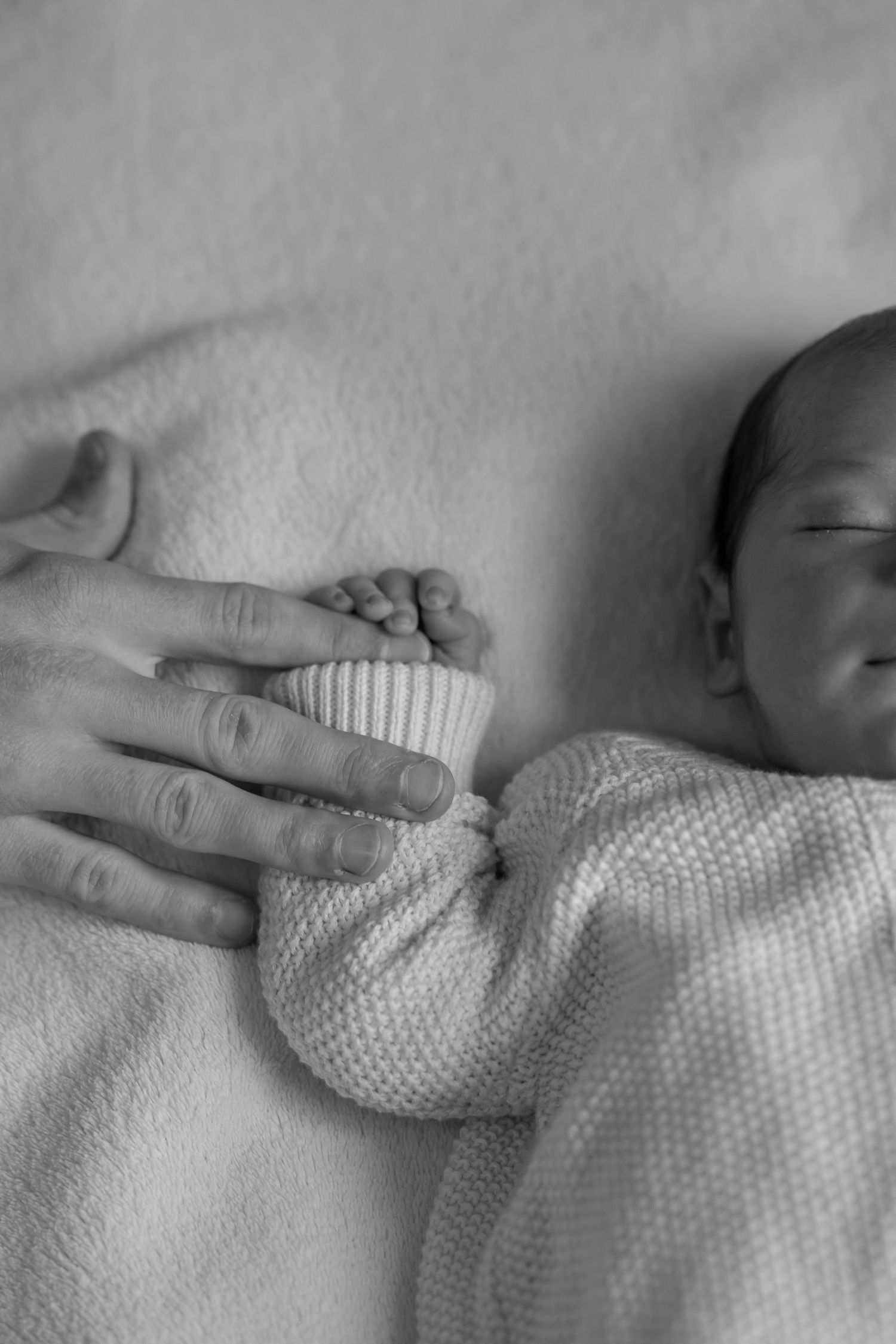 Black and white photo of a newborn baby sleeping while an adult gently holds their tiny hand in a knit sleeve. Newborn Sheffield