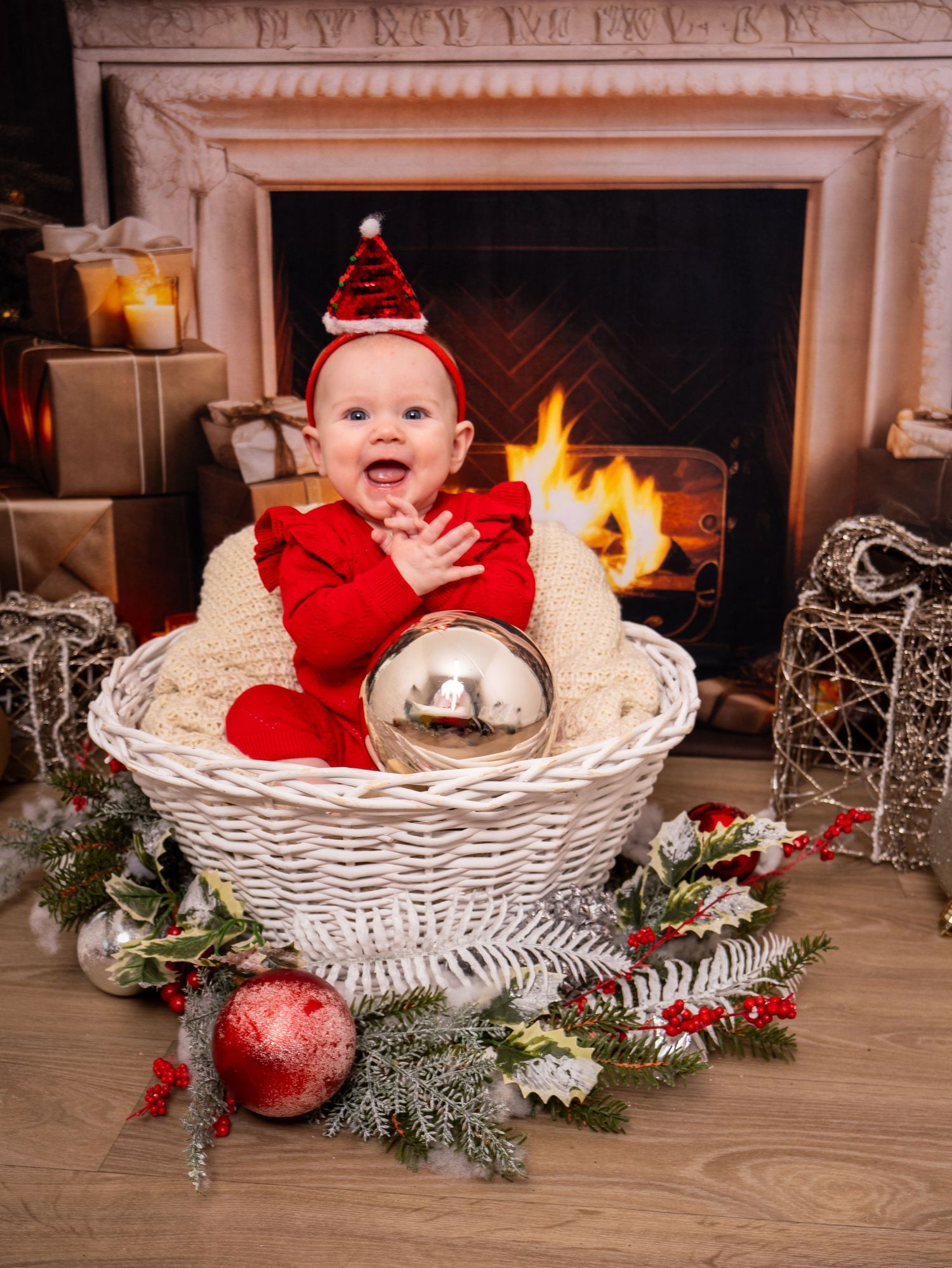 Christmas Photoshoot - A white wicker basket decorated with holiday greenery and red ornaments sits in front of a cozy fireplace.