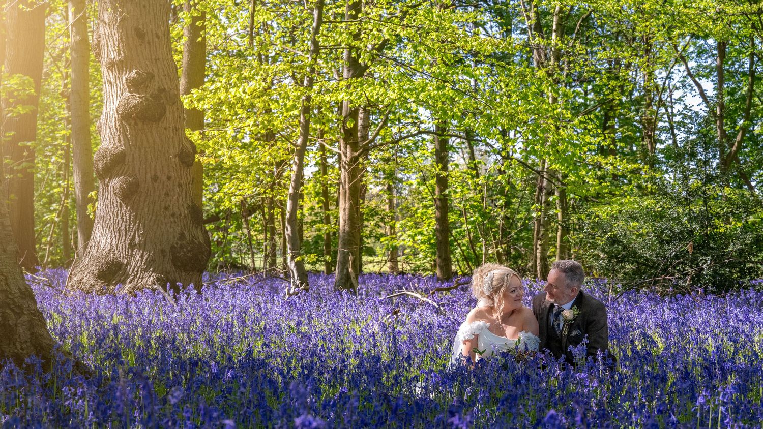 Couple kissing in a stunning bluebell woodland with sunlight filtering through lush green trees.