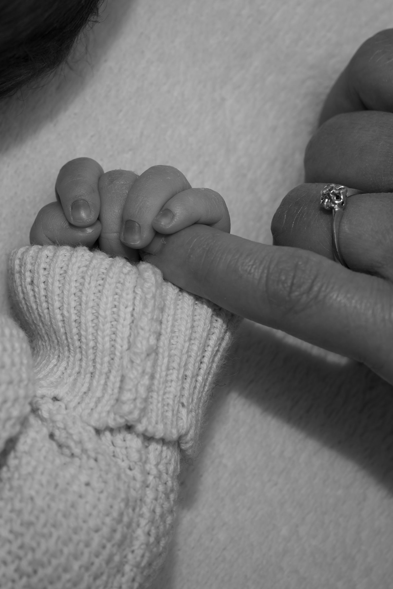 Black and white photo of a newborn baby gripping an adult finger, wearing a knit sweater, with a ring visible on the parent.