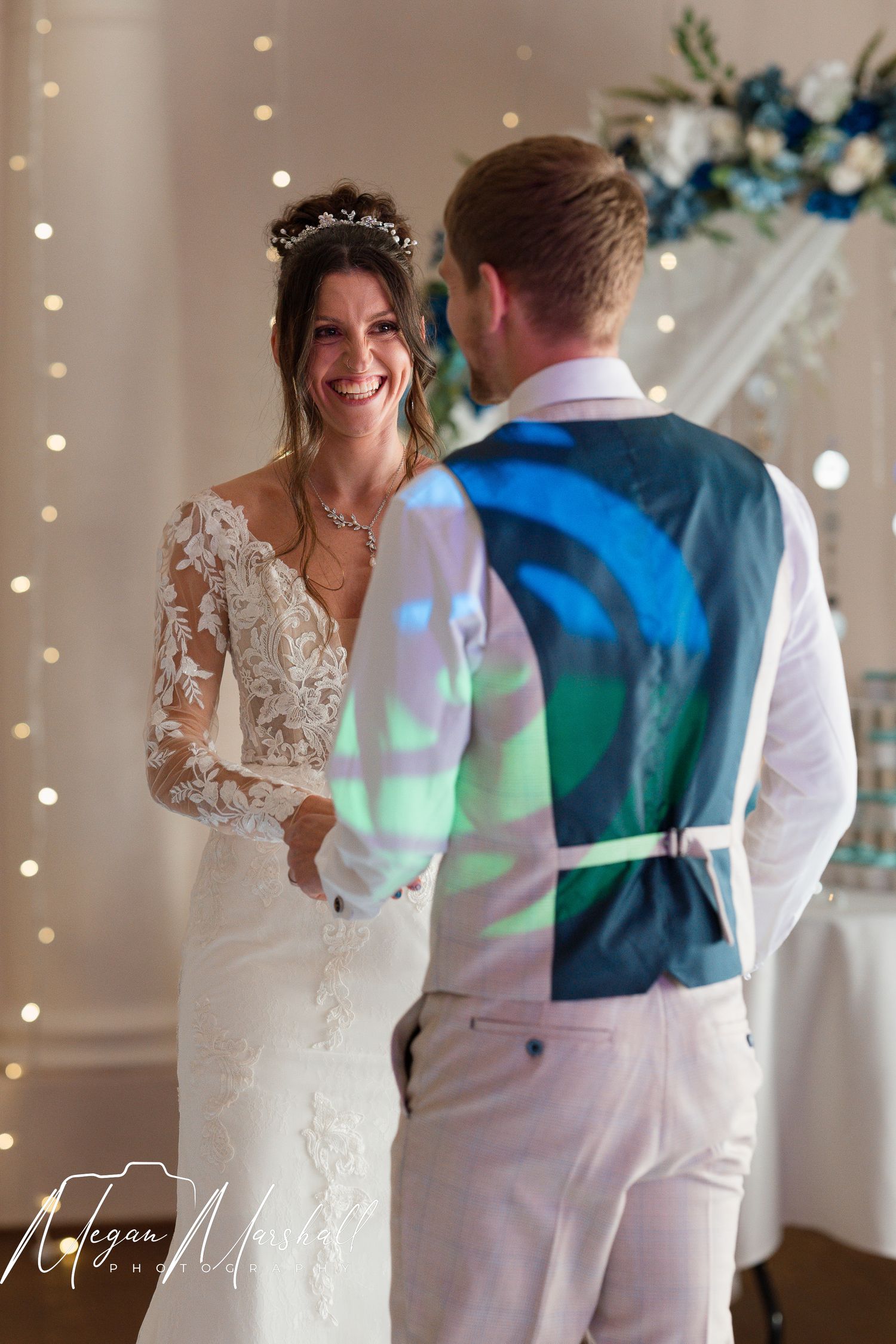 Bride in lace wedding gown smiling at groom during wedding ceremony with fairy lights and floral arch.