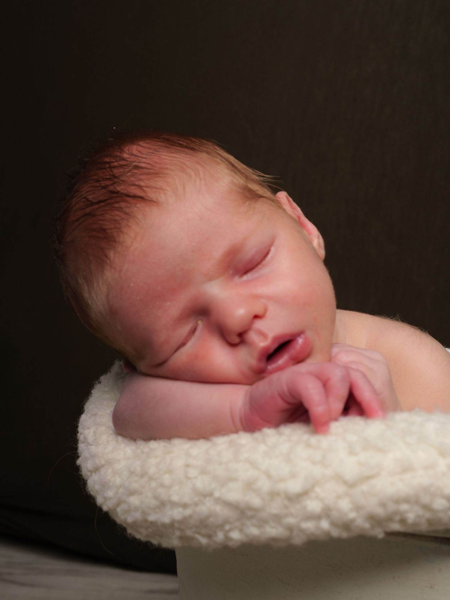 Newborn Photography - A sleeping newborn baby rests peacefully on a soft white textured blanket in a dark setting.
