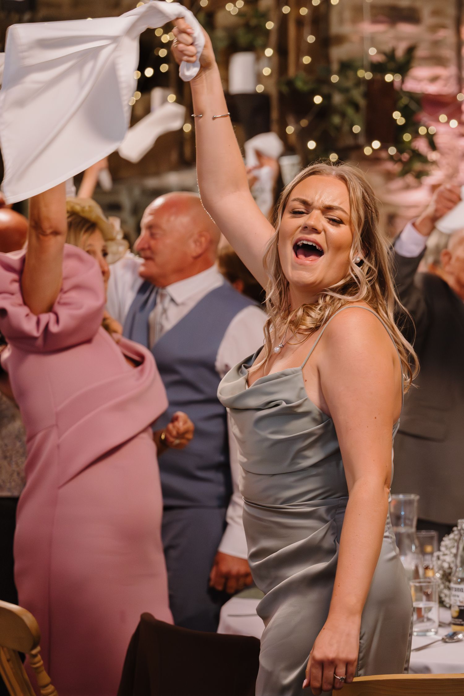 Joyful wedding guest in silver dress raises napkin while celebrating at elegant reception venue.