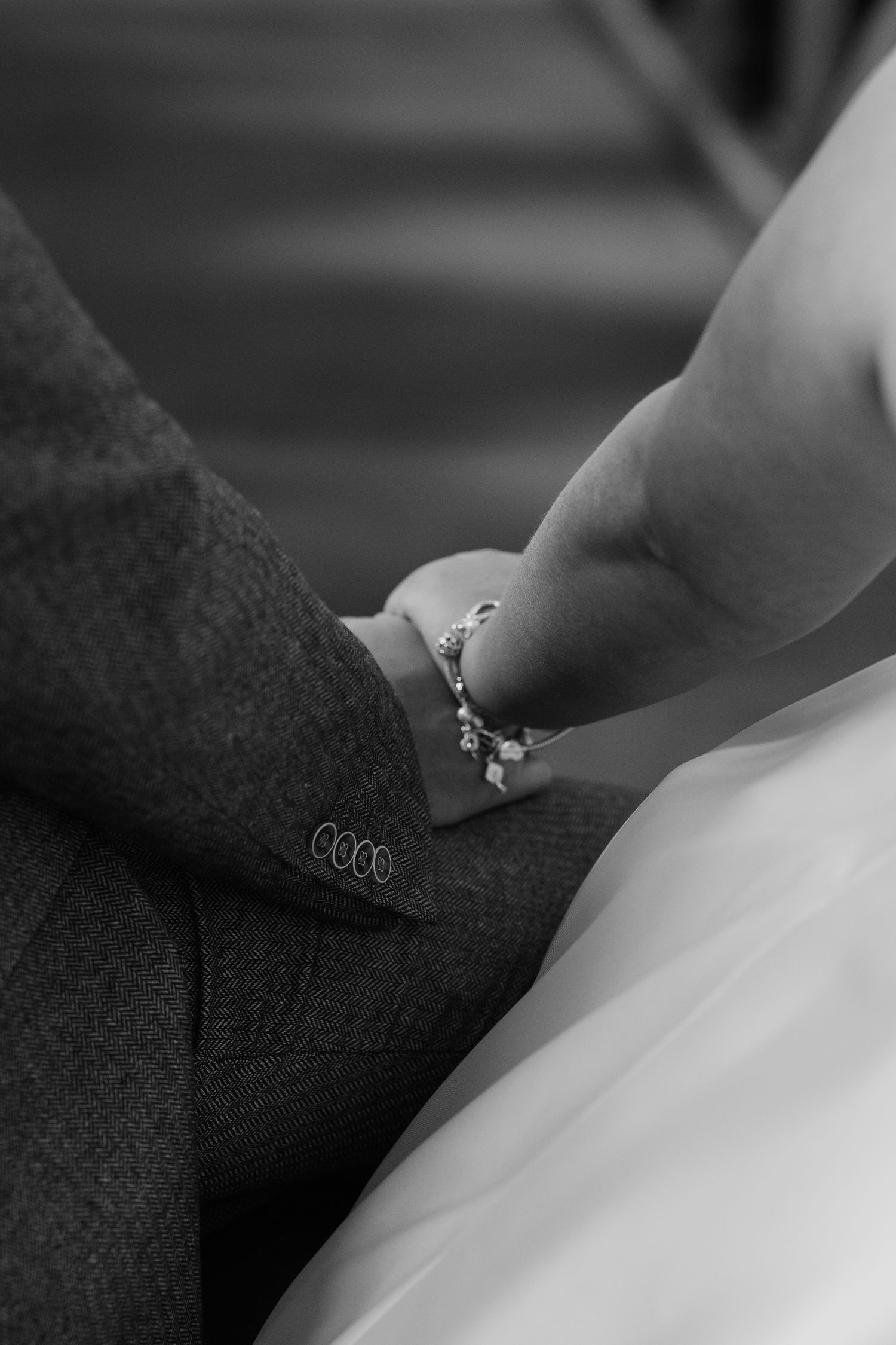 Black and white photo of a bride and groom holding hands, showcasing a delicate bracelet and wedding attire.