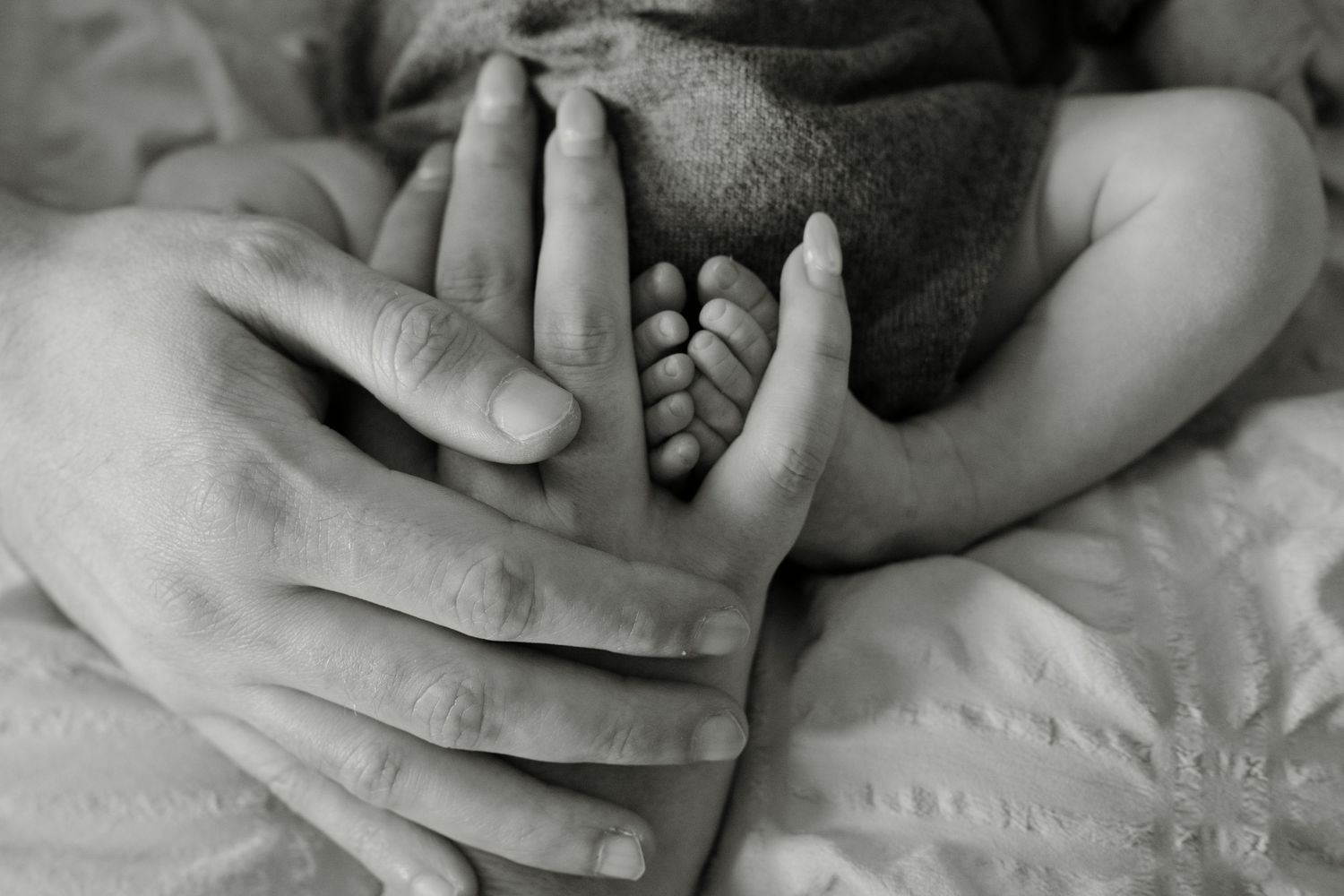 Black and white photo of a parent's hands gently cradling a newborn baby's tiny feet against soft blankets.