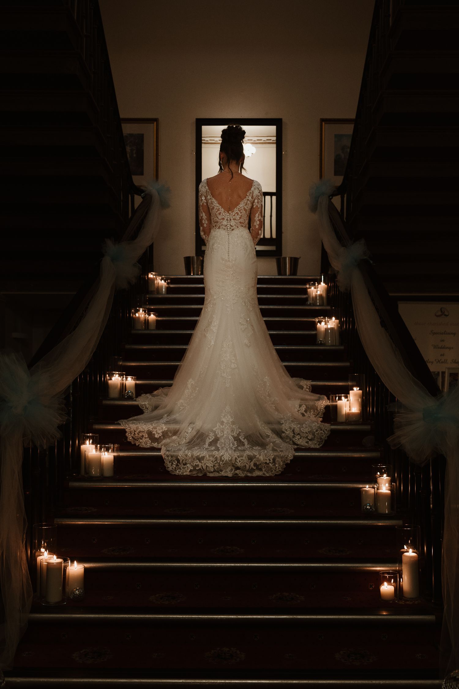 A wedding dress illuminated by candlelight on dark wooden stairs creates a romantic and dramatic silhouette. Venue at Wortley Hall