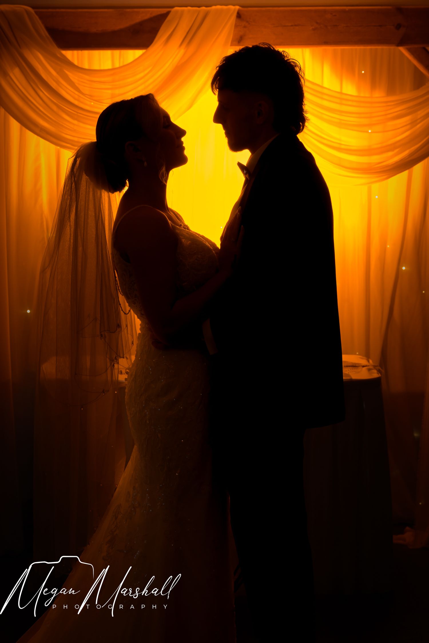 Silhouette of bride and groom sharing romantic moment before golden backlit drape at wedding reception.