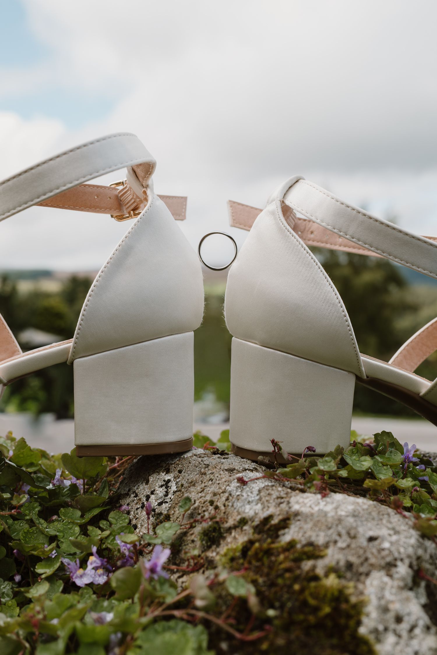 White bridal heels with a gold wedding ring displayed on a mossy rock with purple flowers and green countryside backdrop.
