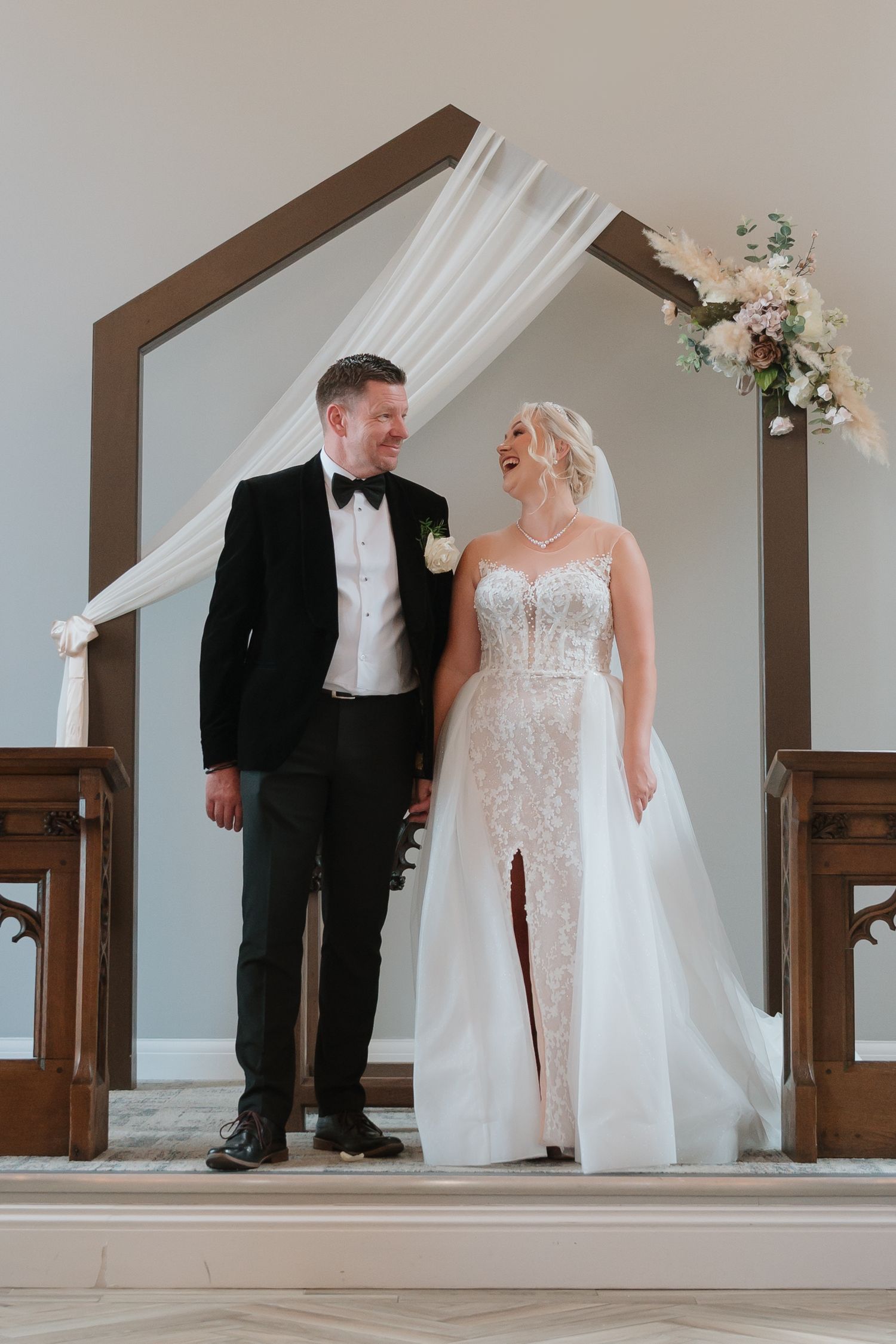 Burntwood Wedding A couple shares a joyful moment at the altar under an elegant arch decorated with white fabric and floral arrangements.