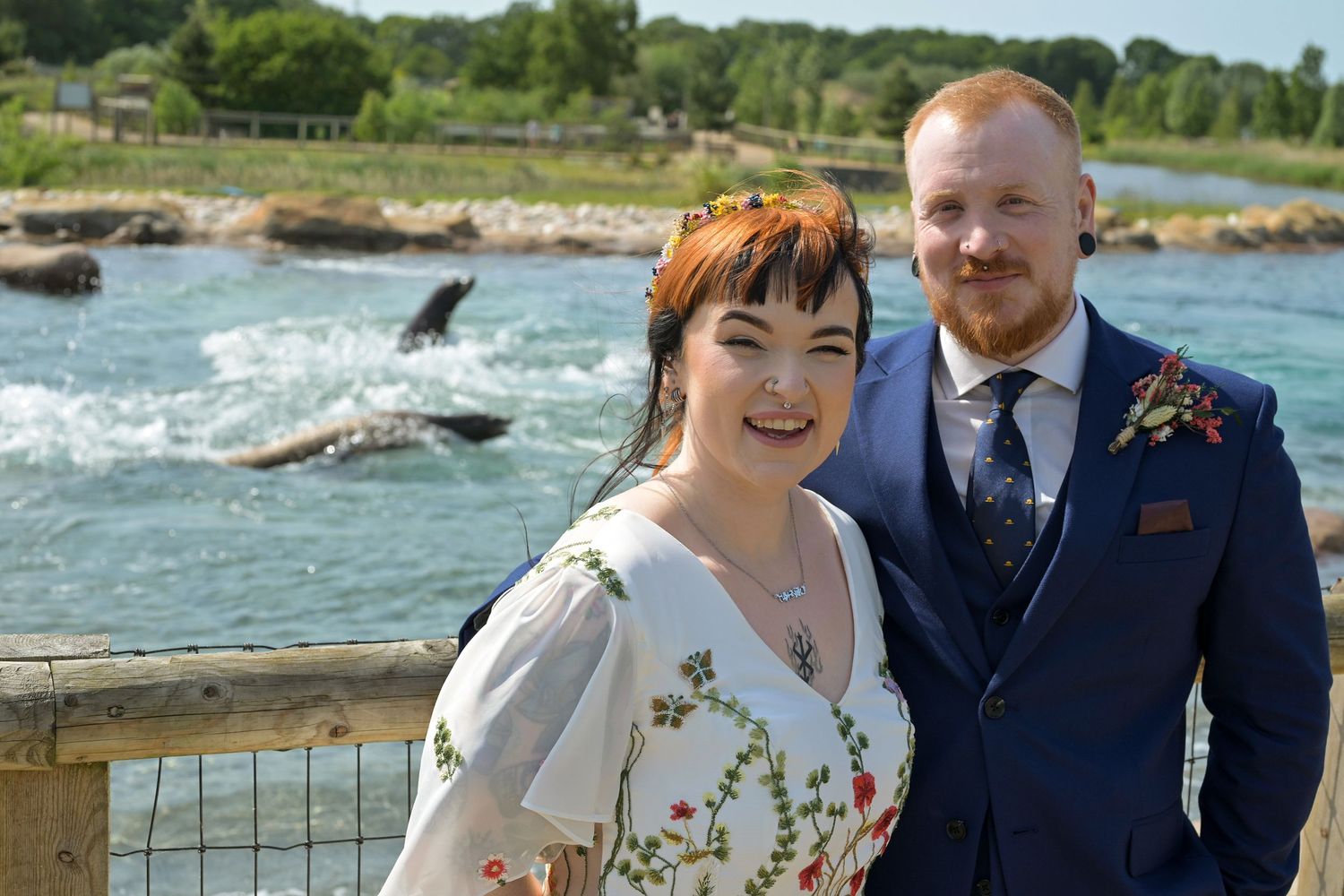 Happy couple posing outdoors near a scenic waterfall on their wedding day, bride in floral dress, groom in navy suit.