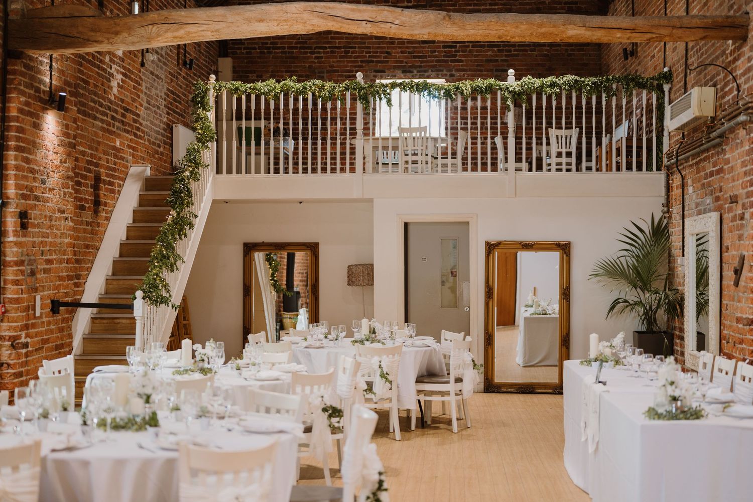 Elegant wedding reception in a rustic barn venue with white tables, brick walls, wooden beams, and lush greenery garland balcony.