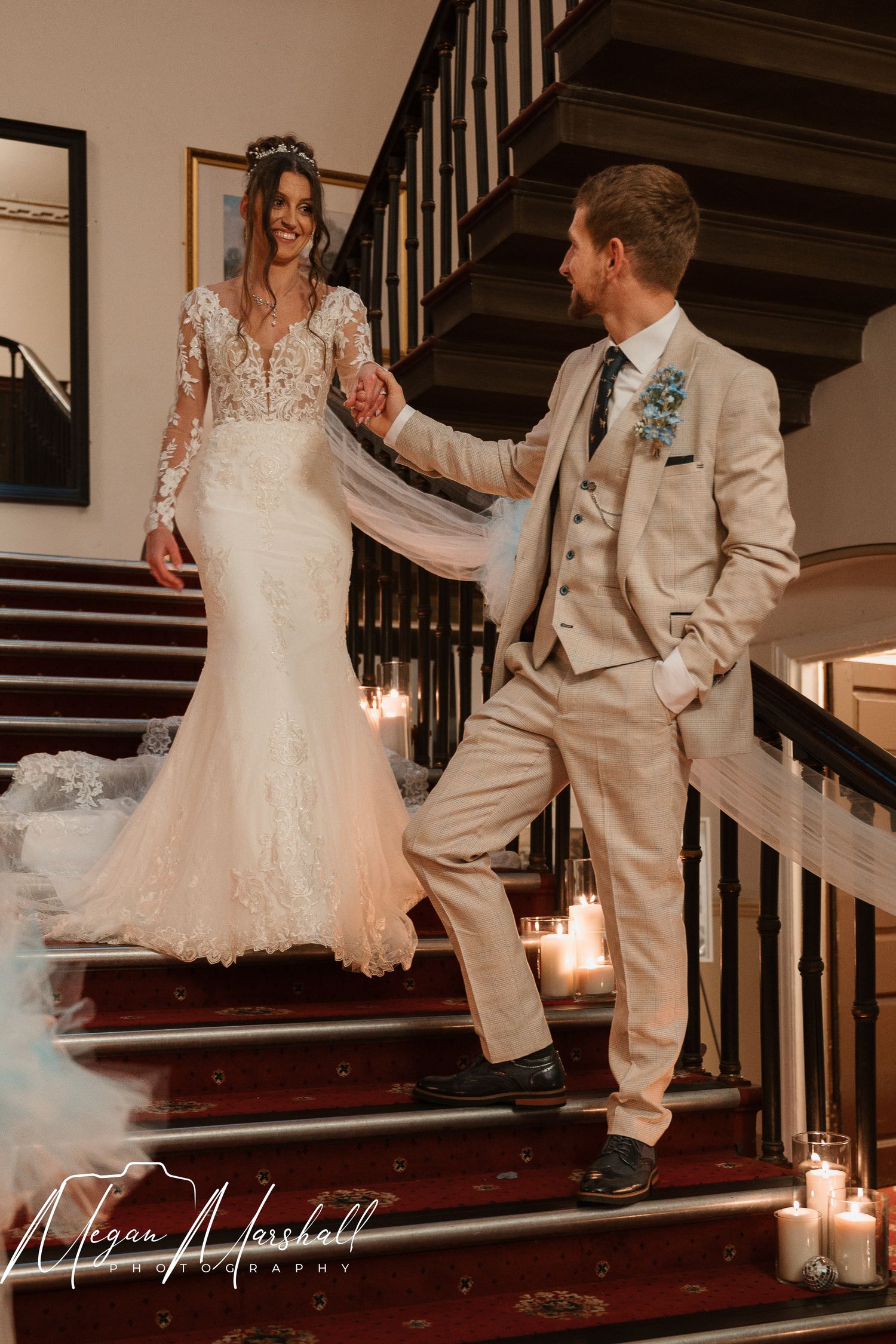 Bride in lace gown and groom in tan suit hold hands on candlelit staircase during elegant wedding portrait.