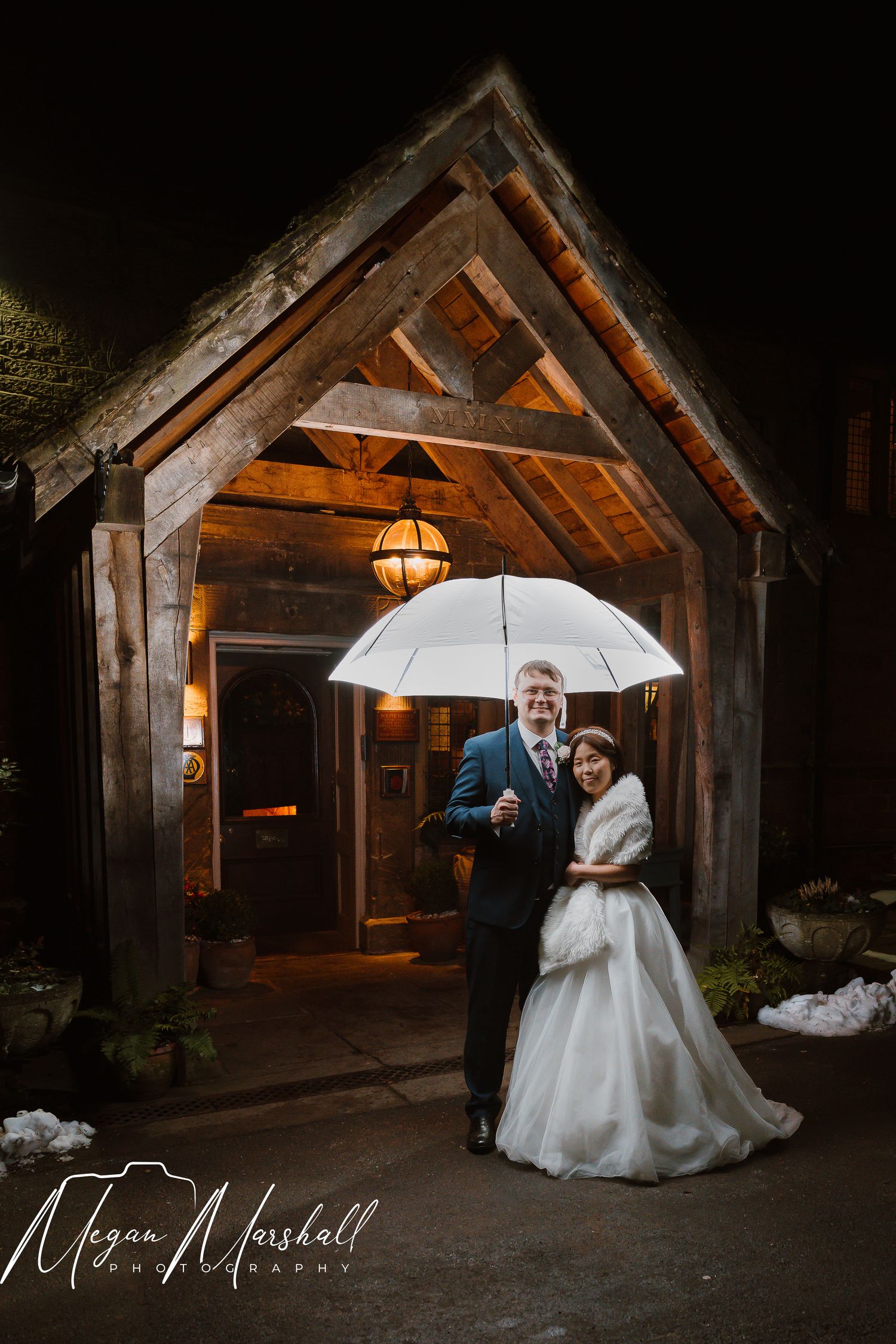 Bride and groom share a white umbrella at night outside a rustic wooden venue entrance, captured by Megan Marshall Photography.