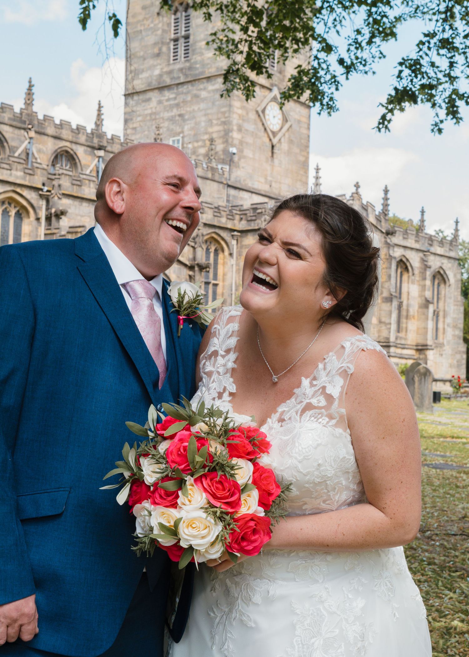 St Marys Church Sheffield - A joyful wedding couple shares laughter while holding a vibrant red and white bouquet outside a church.