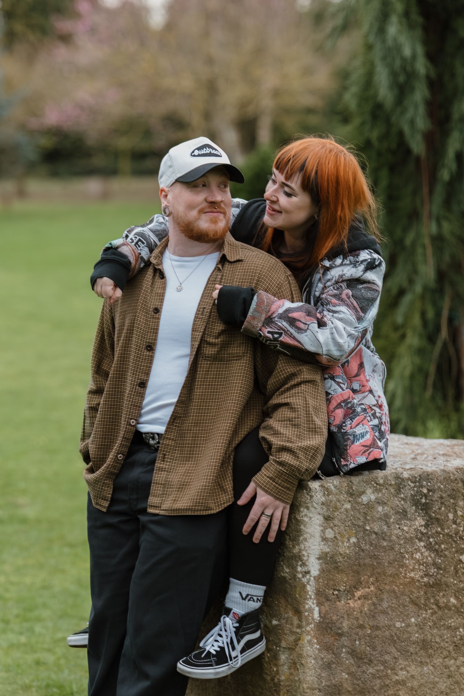 Couple posing outdoors in a park, man in brown shirt and cap, woman with red hair in patterned jacket leaning on stone wall.