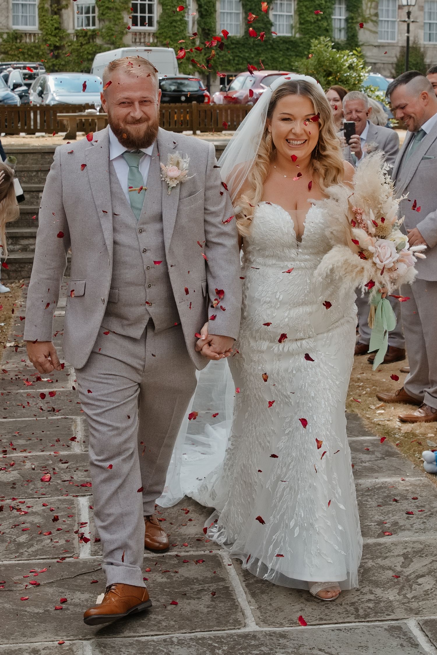 Newlywed couple walking through confetti shower, bride in white lace gown holding bouquet, groom in beige suit.