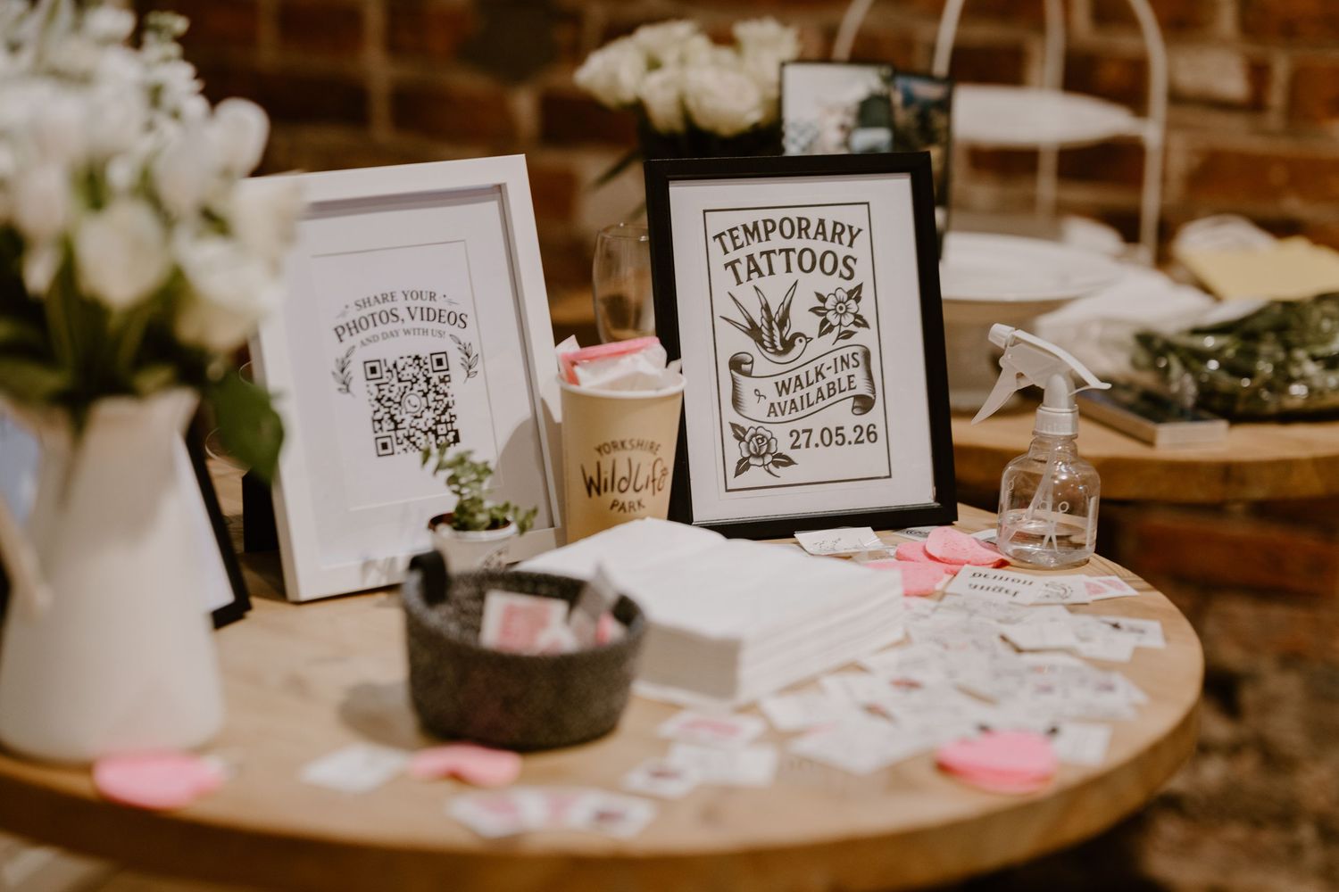 A wedding temporary tattoo station on a round wooden table with framed signs, scattered tattoos, and floral arrangements.