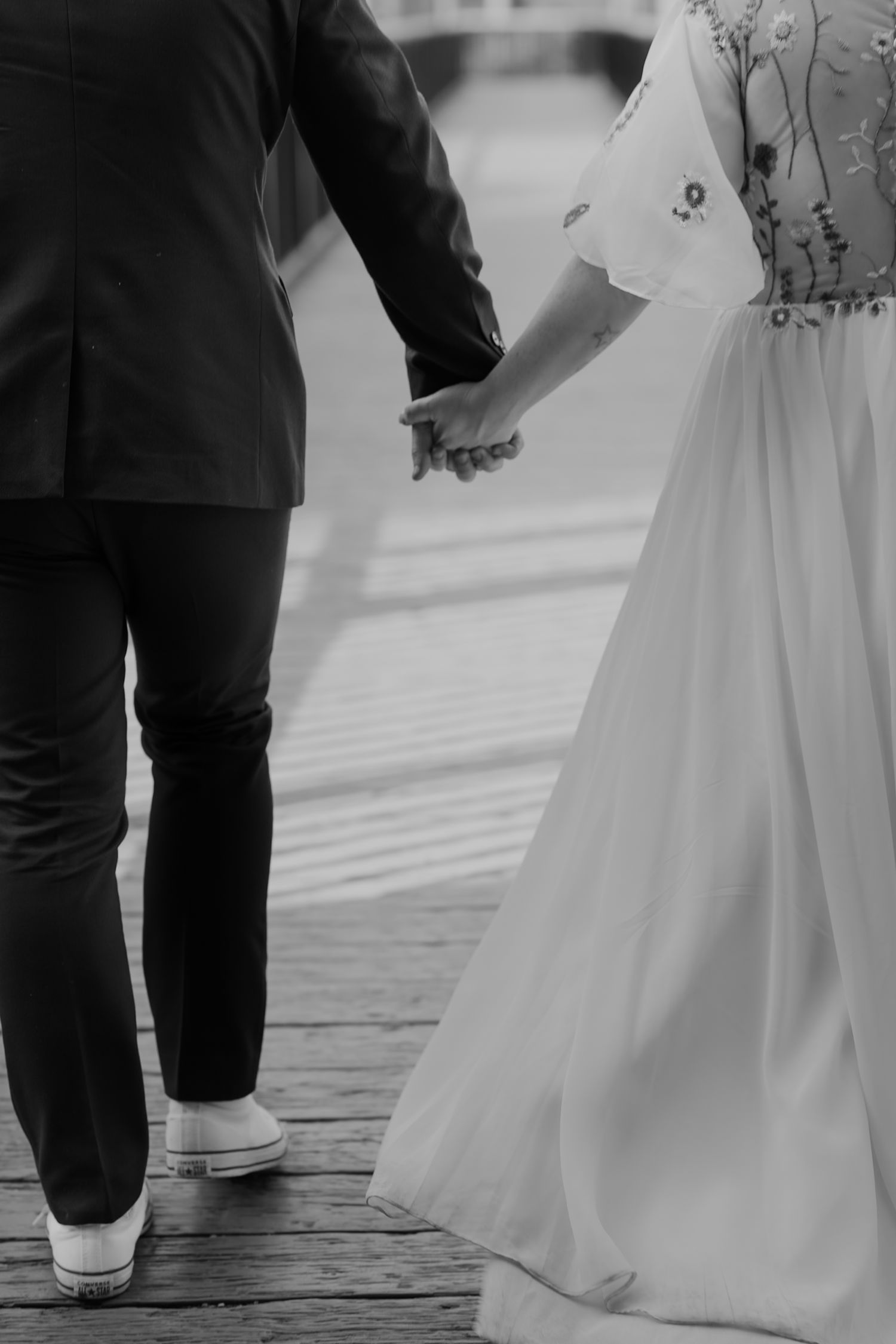 Black and white photo of a bride and groom holding hands while walking on a wooden dock near water.