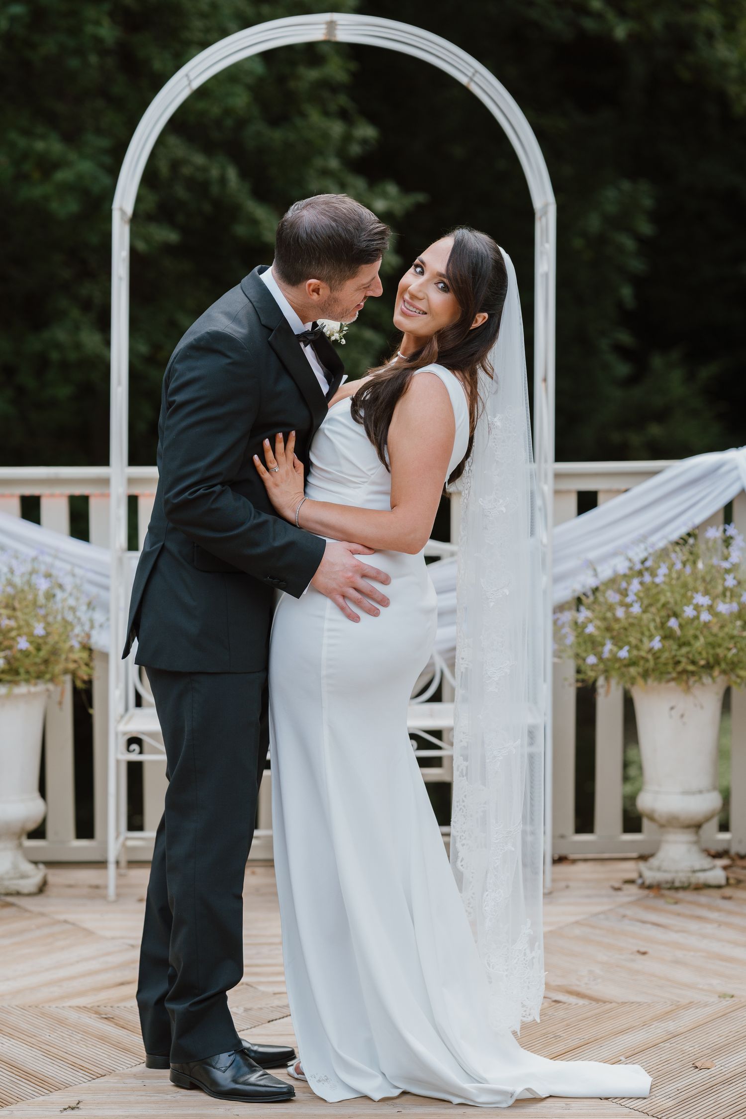 A bride and groom embrace romantically under a white arch on an outdoor deck, surrounded by lush greenery.