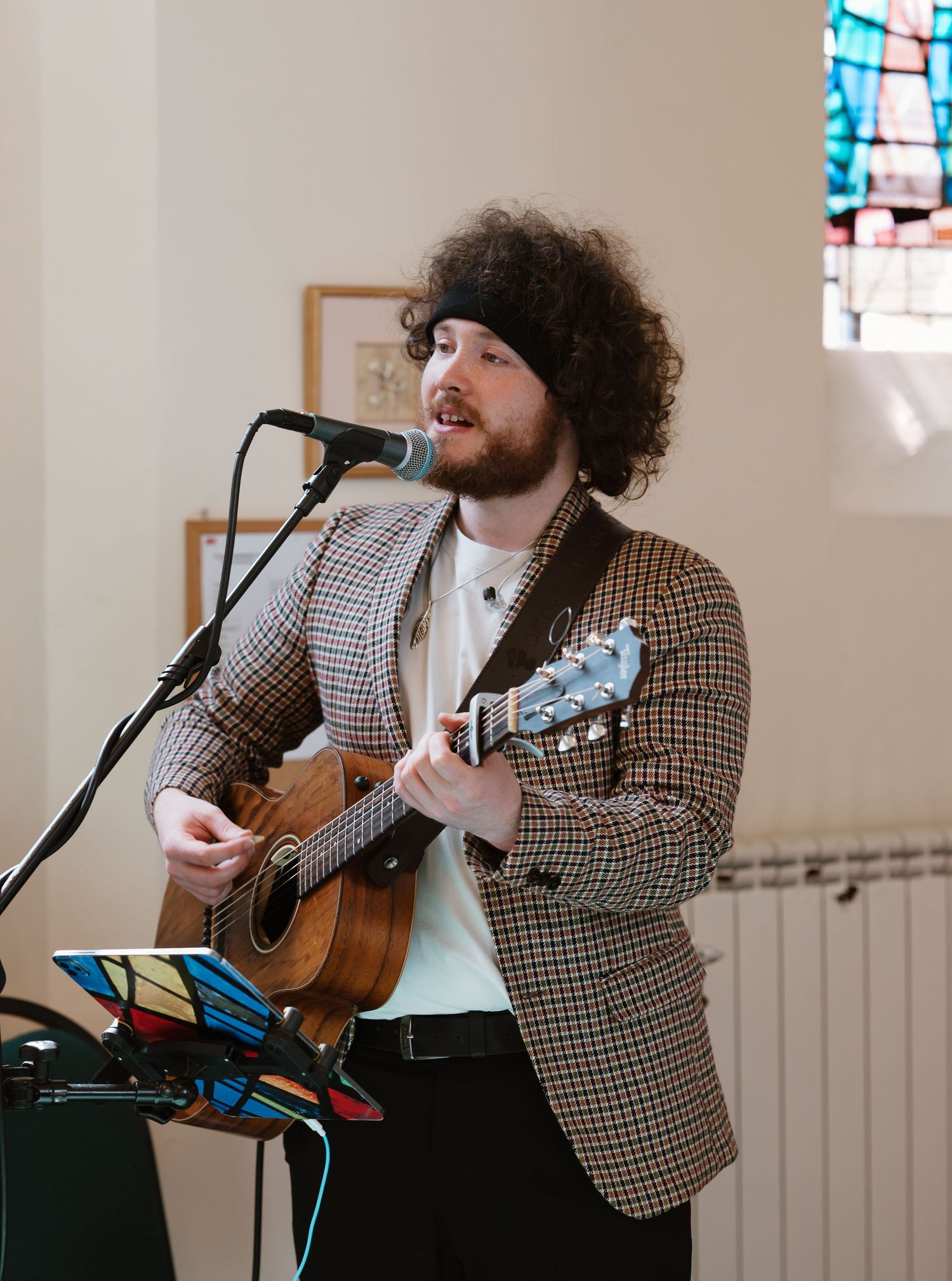 Curly-haired musician playing ukulele and singing into microphone in a church venue wearing a checkered blazer.