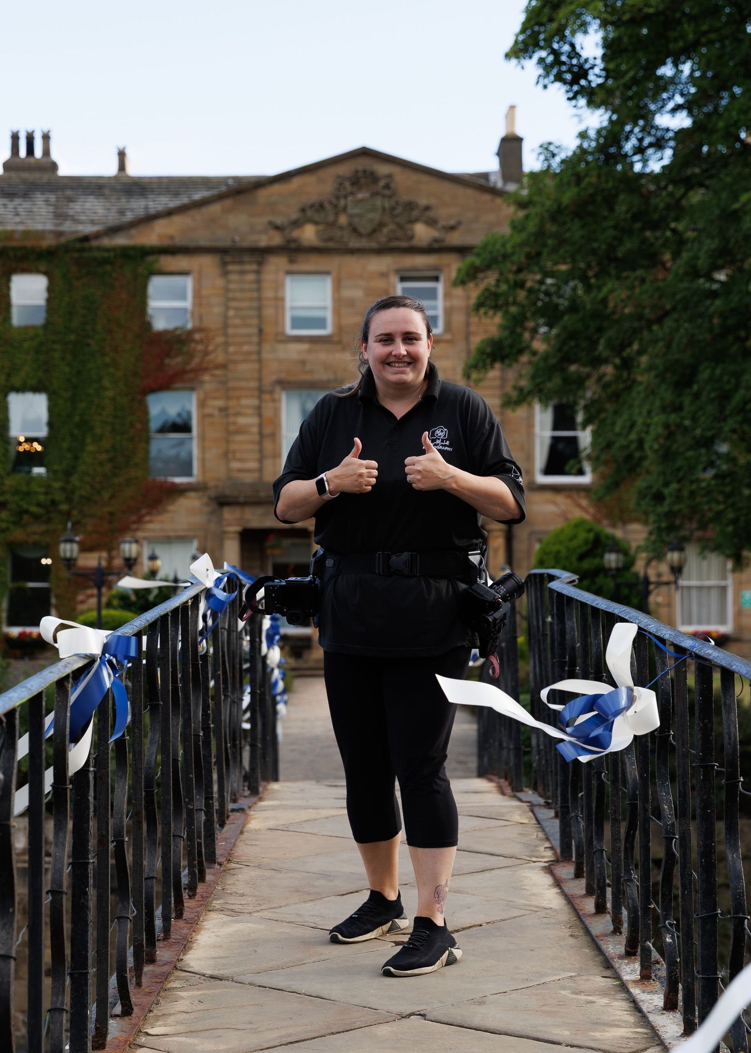 Woman in black uniform giving thumbs up on a bridge outside a grand stone building decorated with blue and white ribbons.