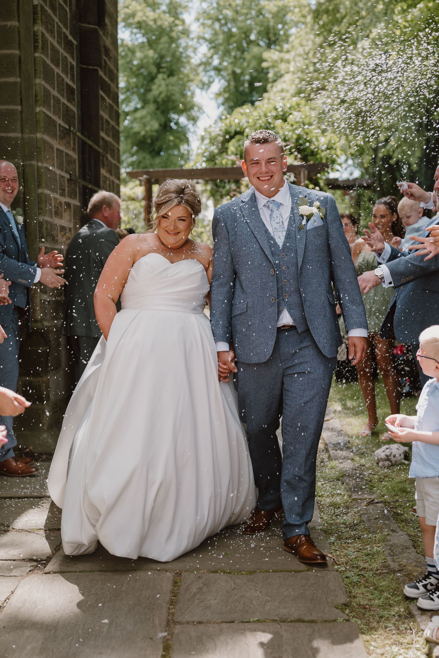 Bride and groom walk through confetti shower outside church on sunny wedding day.