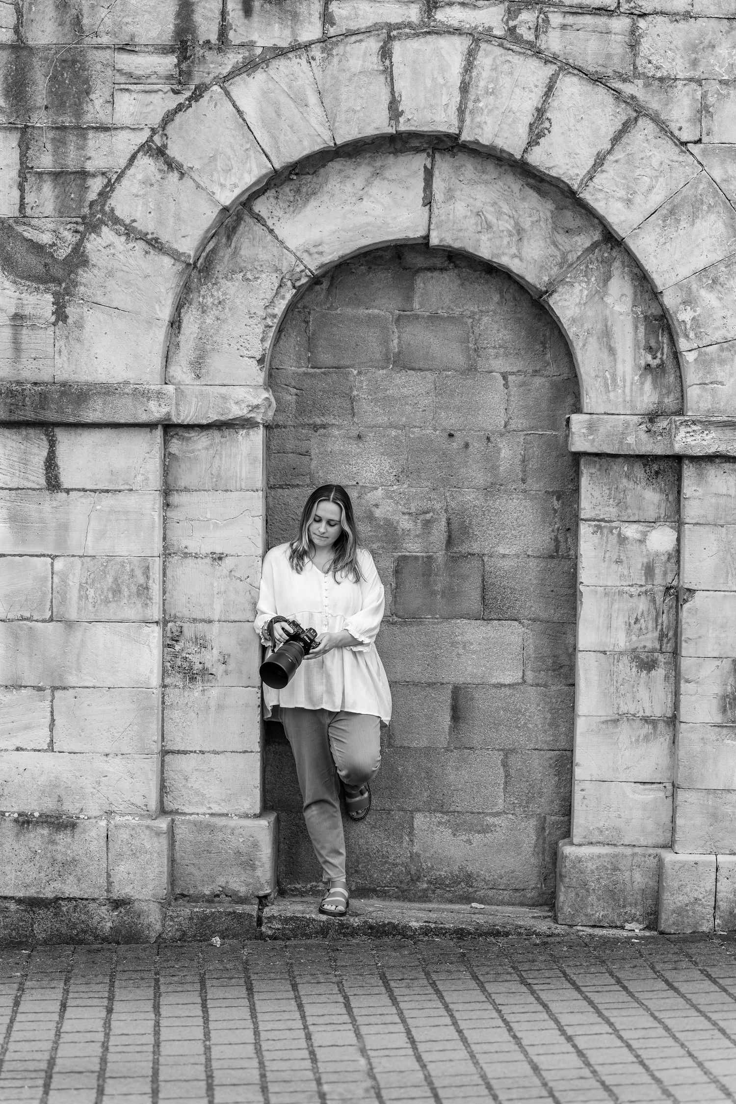 Black and white photo of a woman leaning against a stone arch doorway holding a camera.