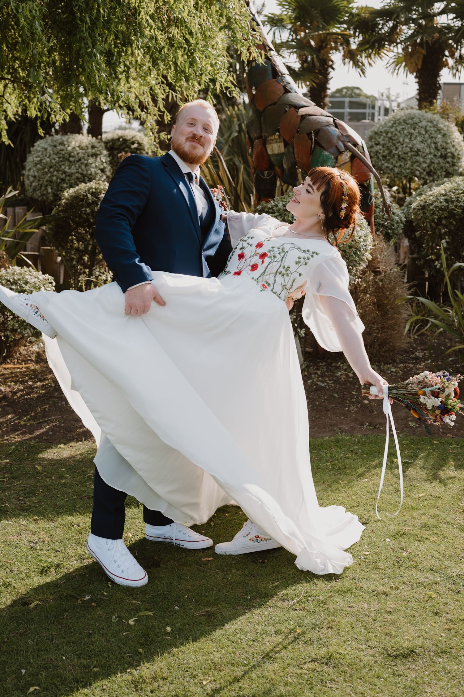 Bride and groom laughing in garden, groom lifting bride's white floral wedding dress, wearing navy suit and white sneakers.
