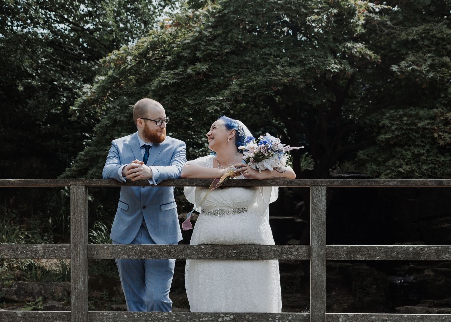 A bride and groom share a romantic moment on a wooden bridge surrounded by lush green trees on their wedding day.