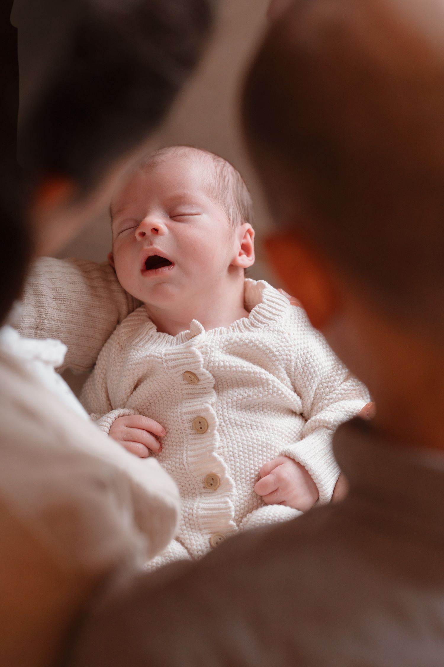 Newborn baby in white knit onesie yawning while being held by parents in a warm, intimate setting.